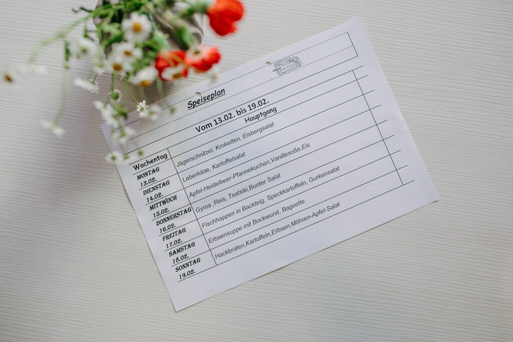 Ein Speiseplan liegt auf einem weißen Tisch neben einem kleinen Strauß mit weißen und roten Blumen. Der Speiseplan zeigt die Hauptgerichte für eine Woche vom 13.02. bis 19.02. Aufgelistet sind verschiedene Gerichte wie Jägerschnitzel, Leberkäse, Apfel-Heidelbeer-Pfannkuchen, Gyros, Fischschnuppen, Erbsensuppe und Hackbraten, jeweils mit Beilagen wie Kartoffelsalat, Reis, Gemüse oder Salat. Die Tabelle ist ordentlich gedruckt und trägt oben den Titel „Speiseplan“. Die Szene wirkt einladend und organisiert.