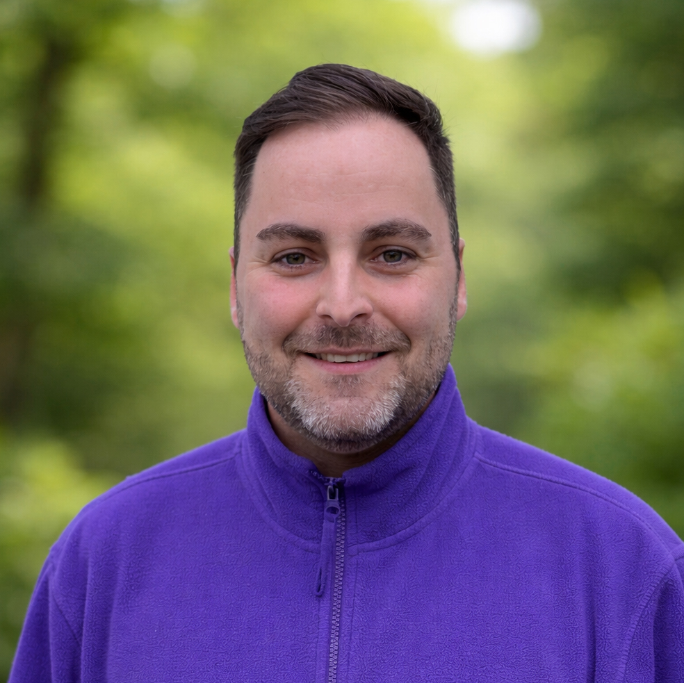 Mitarbeiter eines Pflegedienstes in violetter Arbeitsjacke, Porträtaufnahme im Außenbereich mit natürlichem Hintergrund.