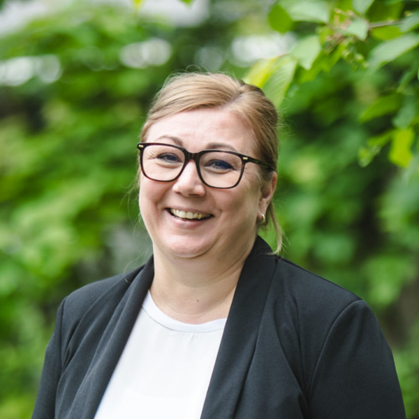 Lächelnde Frau mit blonden, zurückgebundenen Haaren und schwarzer Brille, trägt einen schwarzen Blazer und weißes Oberteil, steht vor grünen Bäumen im Freien.